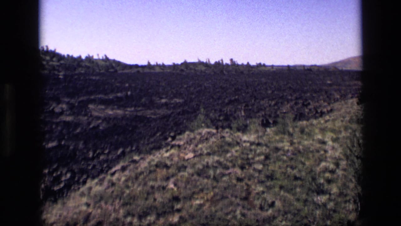 1972 Idaho 8mm frame showing dark rocky terrain, scattered shrubs, dry grasses, and low hills under a pale blue sky.