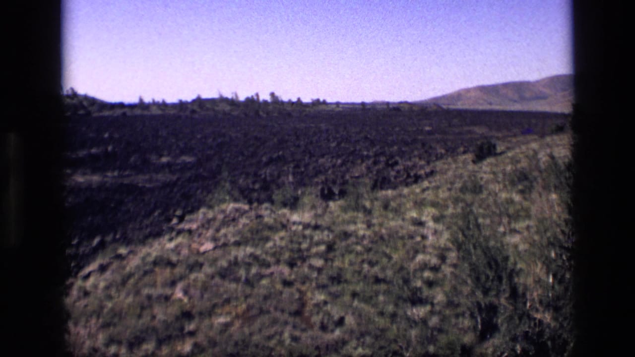 1972 Idaho home-movie frame with dark rocky ground, scrub vegetation, a grassy foreground edge, and rolling hills in the distance.
