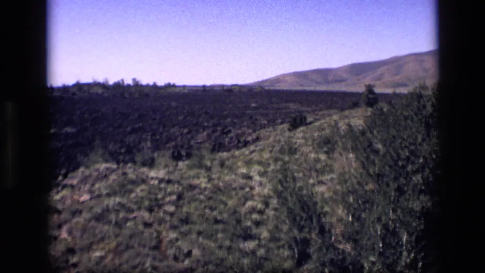 1972 Idaho 8mm frame showing a rugged dark field, green shrubs, dry grass, and a broad rounded hill beyond the plain.