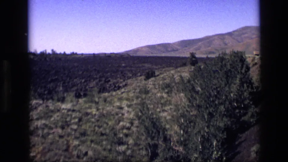 1972 Idaho frame from an 8mm pan, with scrubby foreground plants, dark rocky plain, and larger hills beyond the open terrain.