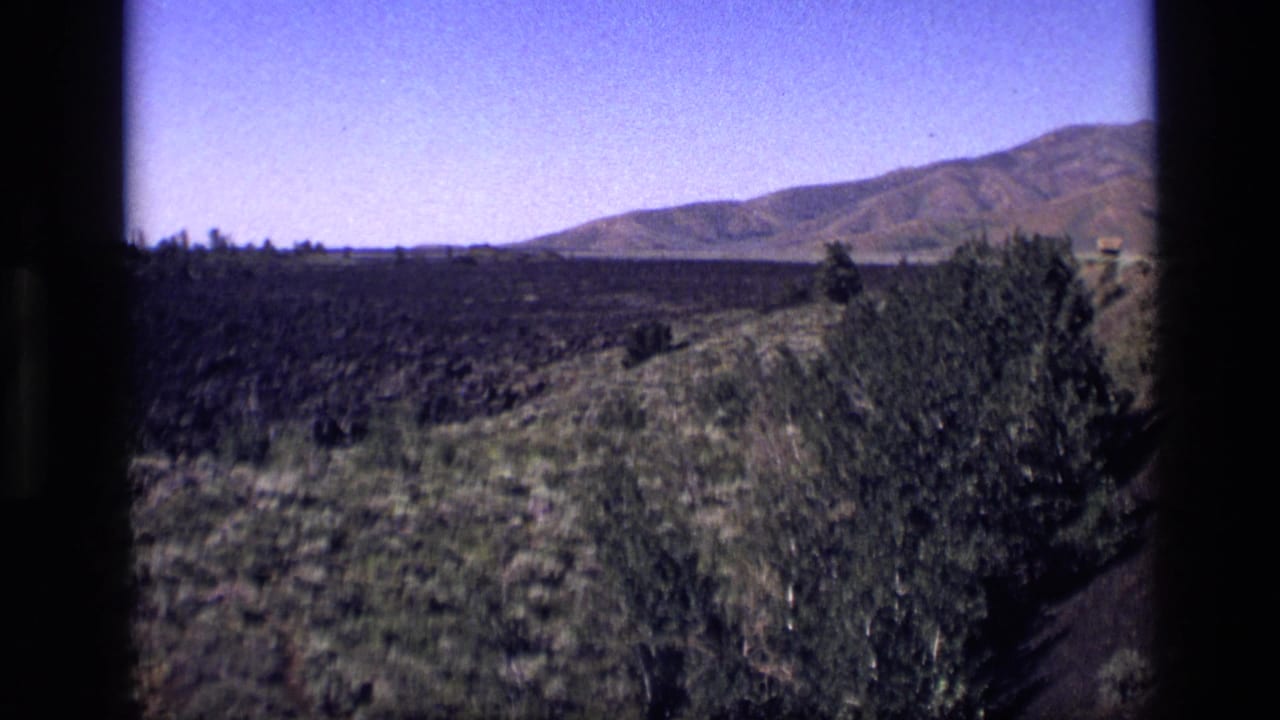 Dry hillside landscape with shrubs, distant low mountains, blue sky, and dark film borders.