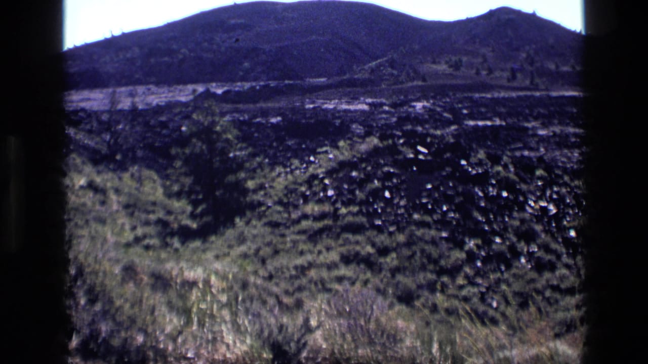 Open hillside with dark rocky ground, grasses, distant mountain ridge, and black frame edges.