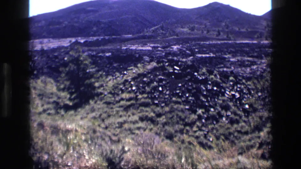 1972 Idaho 8mm frame with dry foreground grass, dark uneven terrain, and rounded mountain slopes in the distance.