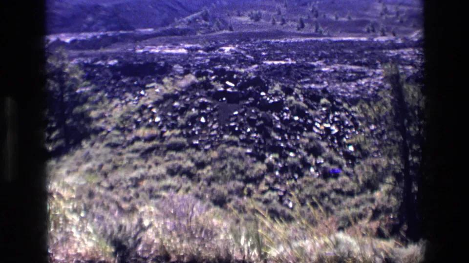 1972 Idaho landscape frame showing dark textured ground, dry grasses, shrubs, and a distant hillside within 8mm borders.