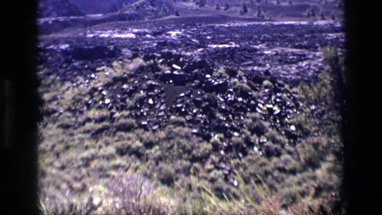 1972 Idaho 8mm frame showing uneven rocky terrain, scrub vegetation, and bright outdoor light across a rugged natural area.