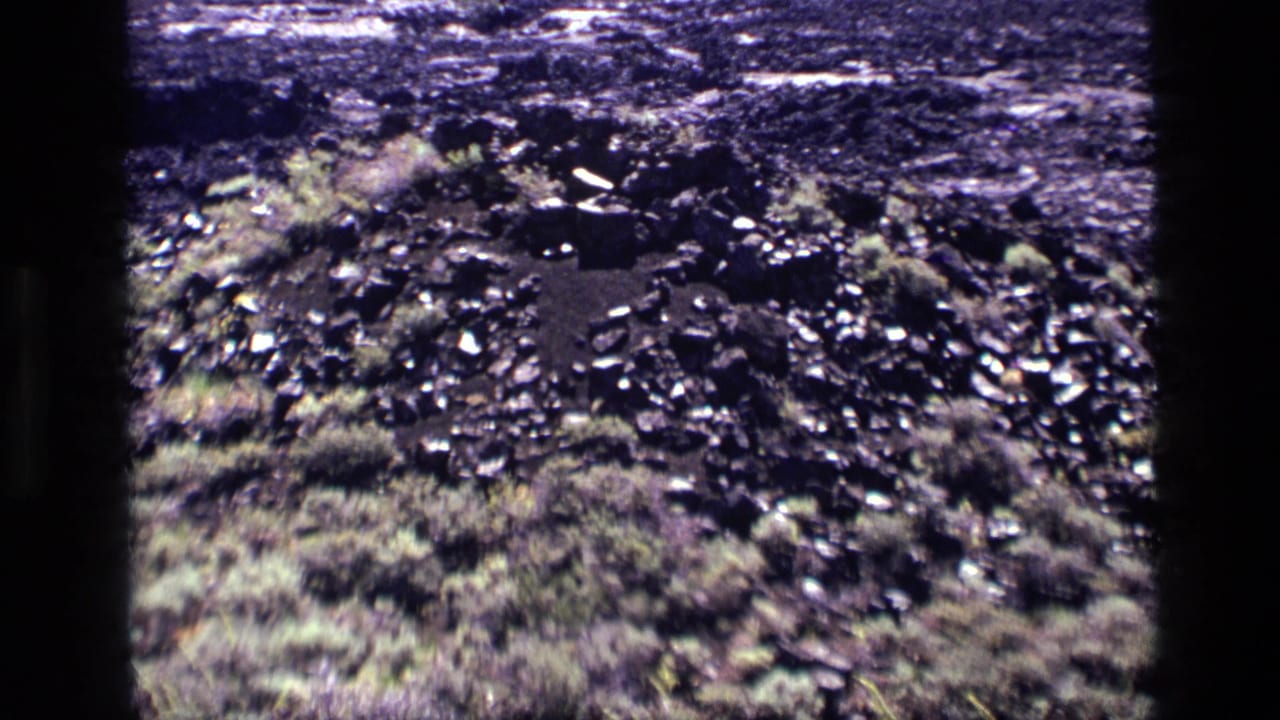 1972 Idaho home-movie frame with dark rocky debris, low vegetation, and a slightly closer handheld view of the terrain.