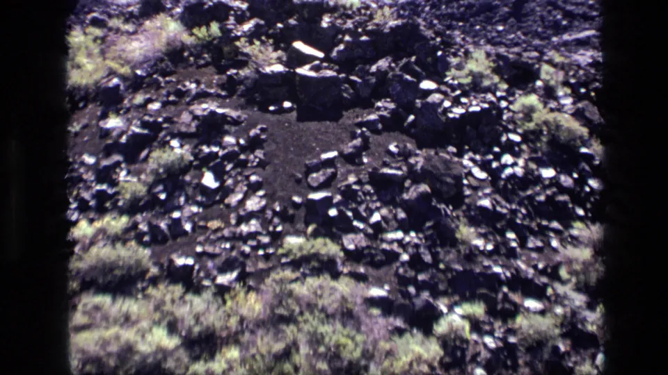 1972 Idaho 8mm frame showing a circular bare-soil depression bordered by dark rocks and sparse vegetation.