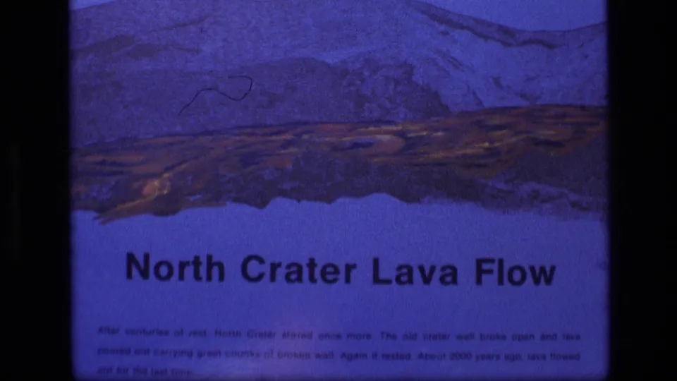 1972 Idaho 8mm frame of the North Crater Lava Flow sign, showing blue-tinted artwork, printed text, and minor film marks.