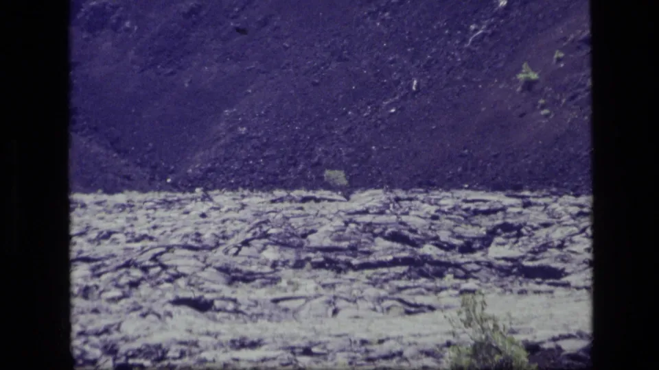 1972 Idaho 8mm frame showing textured North Crater Lava Flow rock with sparse vegetation and a dark slope in the background.