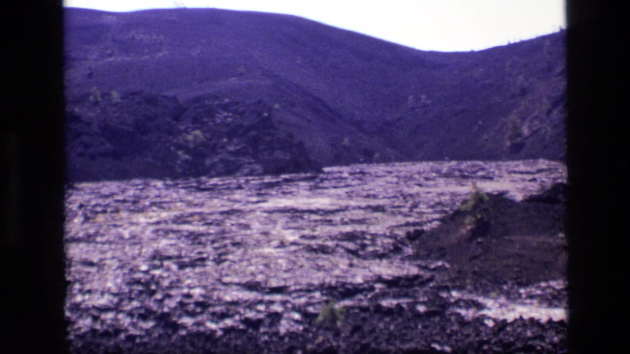 1972 Idaho 8mm frame showing rocky open ground, low grass, distant hills, and a bright sky with strong purple-blue color shift.