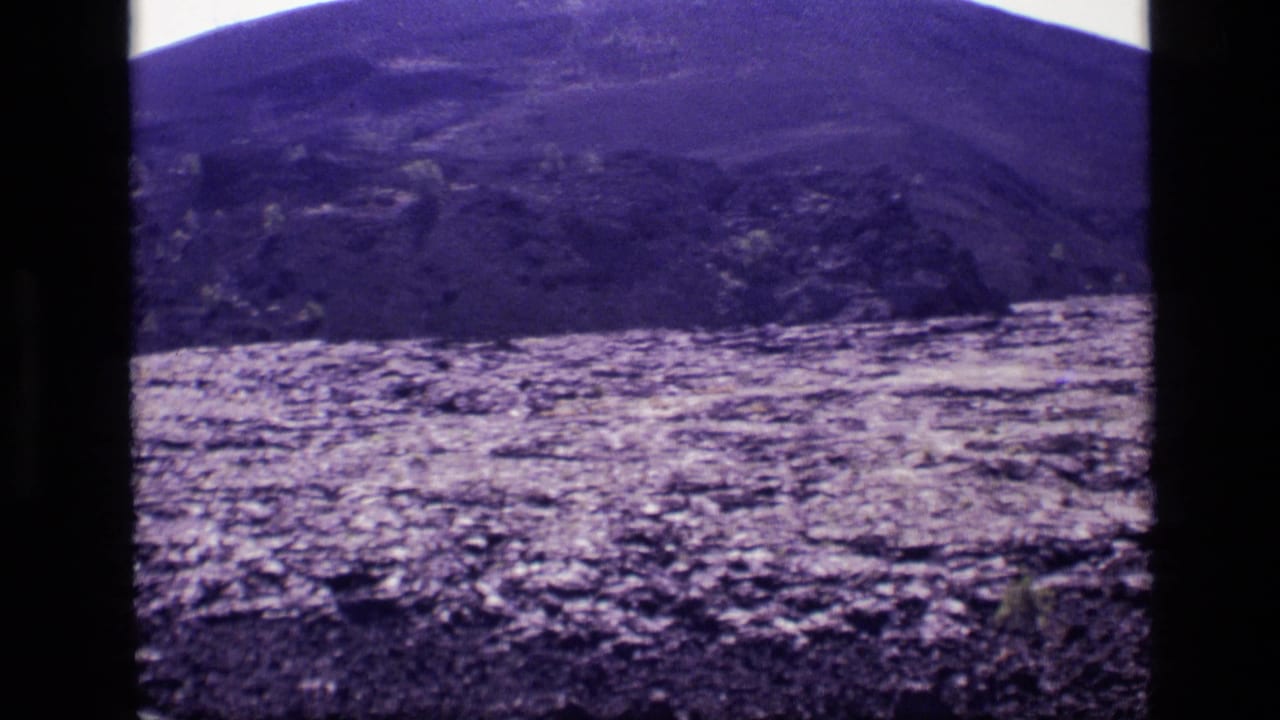 1972 Idaho 8mm frame with rolling hillside, uneven rocky ground, sparse plants, and bright overexposed sky during the camera pan.