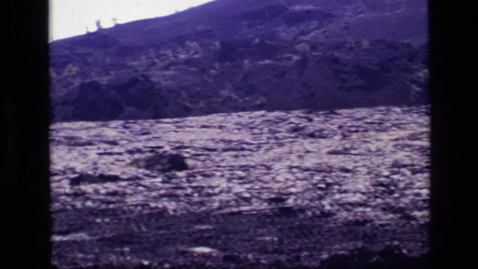 1972 Idaho 8mm frame showing a sloped hillside with rough ground, patches of grass, scattered trees, and a bright sky.