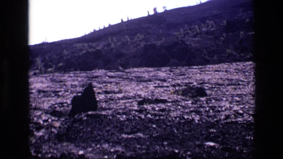 1972 Idaho 8mm frame showing rocky hillside, scattered trees, grass, bright sky, and a small dark human-like silhouette near the foreground.