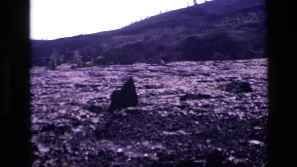 1972 Idaho home-movie frame showing cracked volcanic ground, a foreground rock form, low hills, and bright sky.