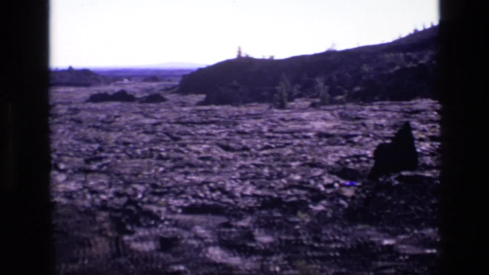 1972 Idaho landscape frame with barren volcanic ground, scattered brush, low hills, and a foreground rock shape.