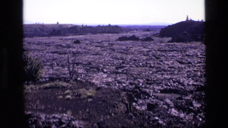 1972 Idaho 8mm frame showing barren cracked terrain, sparse brush, distant low hills, and dark film-frame edges.