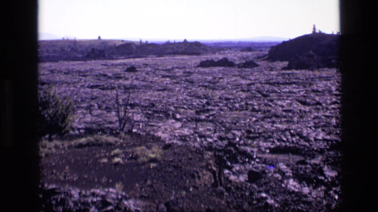 1972 Idaho 8mm frame with rough cracked earth, low horizon, sparse vegetation, and a shadowed rocky mound at the right edge.