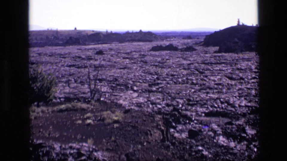 1972 Idaho 8mm frame with rough cracked earth, low horizon, sparse vegetation, and a shadowed rocky mound at the right edge.