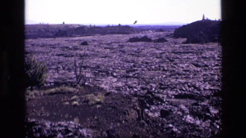 1972 Idaho home-movie frame showing flat rocky wasteland, scrubby vegetation, distant hills, and visible 8mm frame borders.