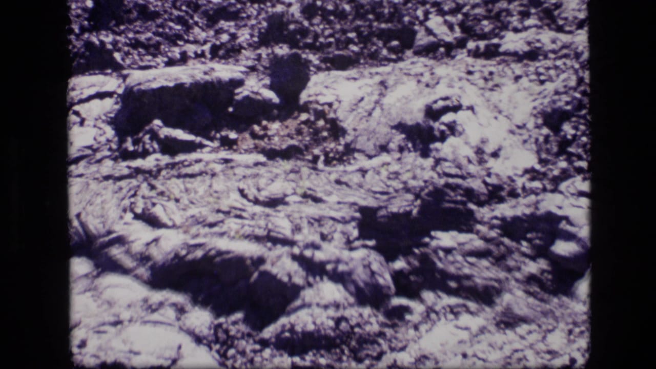 1972 Idaho 8mm close-up of textured volcanic-looking ground, with layered rock, small stones, and high-contrast shadows.