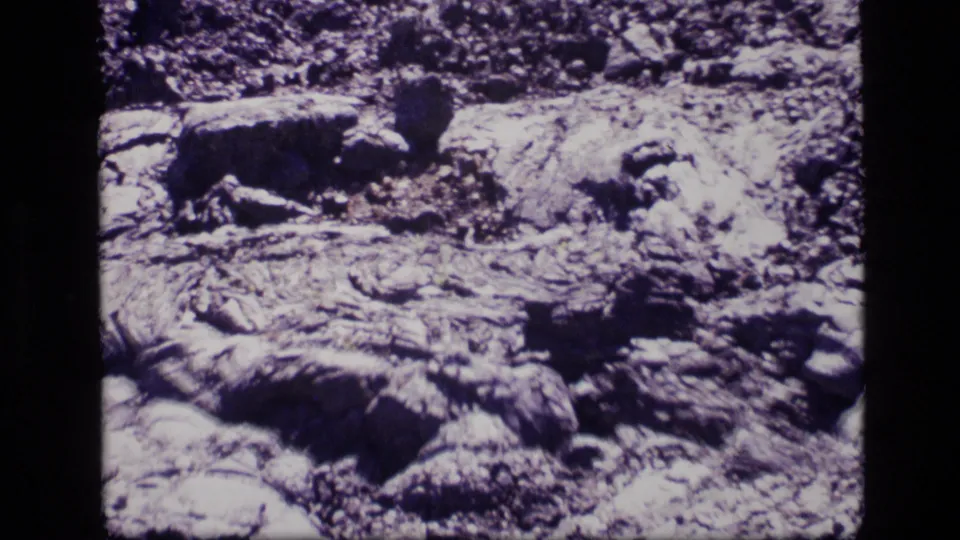 1972 Idaho 8mm close-up of textured volcanic-looking ground, with layered rock, small stones, and high-contrast shadows.