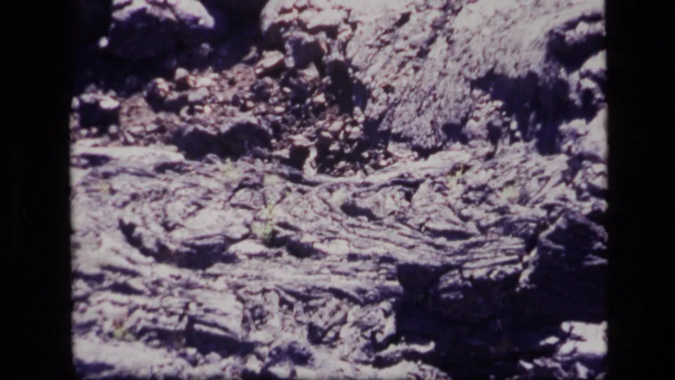 1972 Idaho 8mm close-up of rough volcanic-looking terrain, with pale ridges, gravel fragments, and deep shadowed cracks.