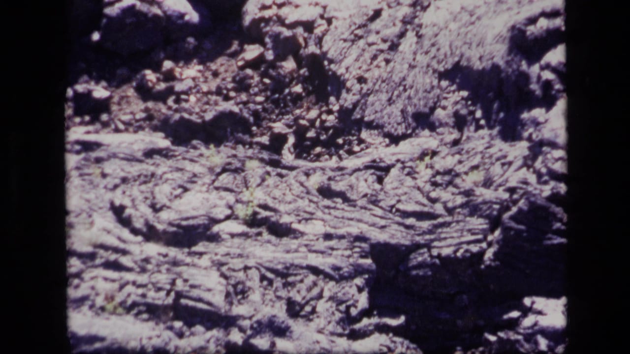 1972 Idaho 8mm frame showing cracked volcanic rock, rough debris, and shadowed edges across a barren lava surface.
