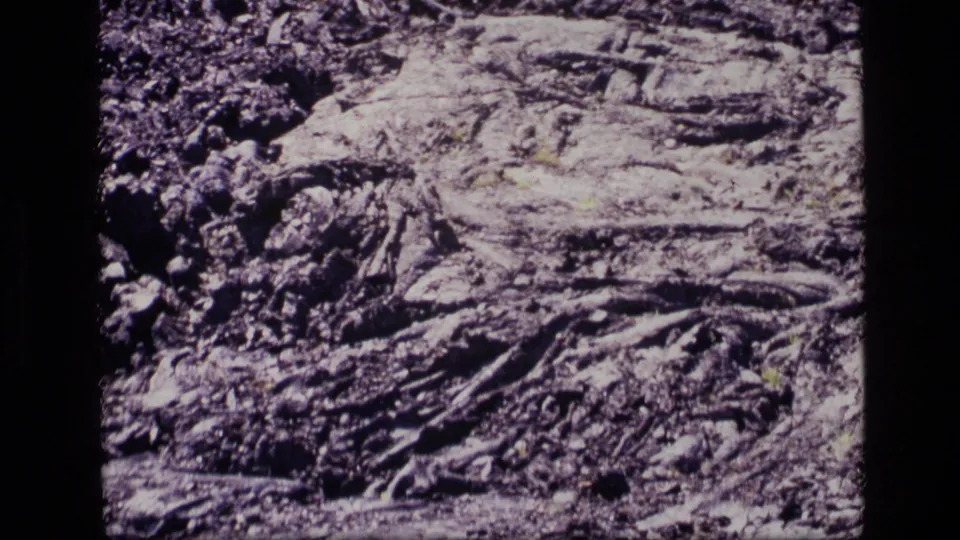 1972 Idaho 8mm frame showing cracked solidified lava, dark volcanic debris, and sparse green growth across rocky terrain.