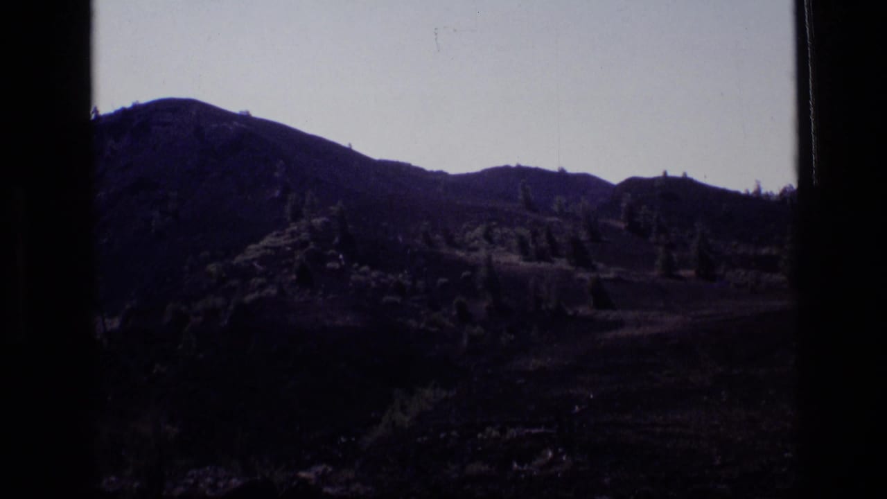 1972 Idaho 8mm view of a shadowed rocky hillside with sparse evergreen trees and a dark mountain ridge beyond.