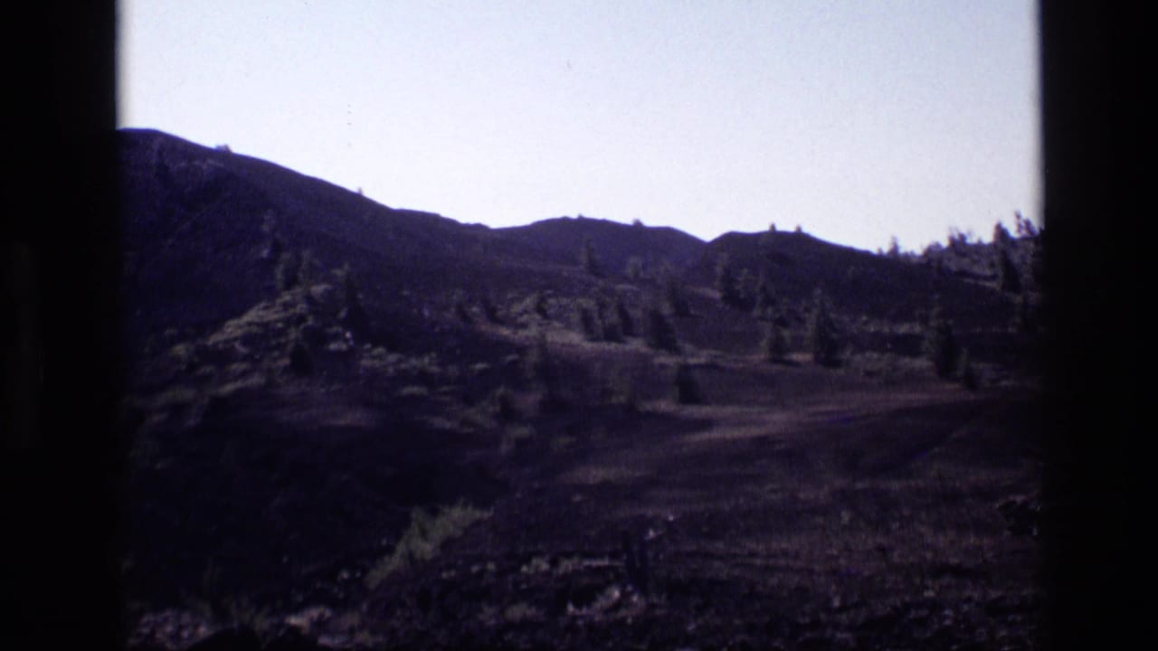 Mountain hillside with dry terrain, evergreen trees, and a pale sky framed by dark film edges.