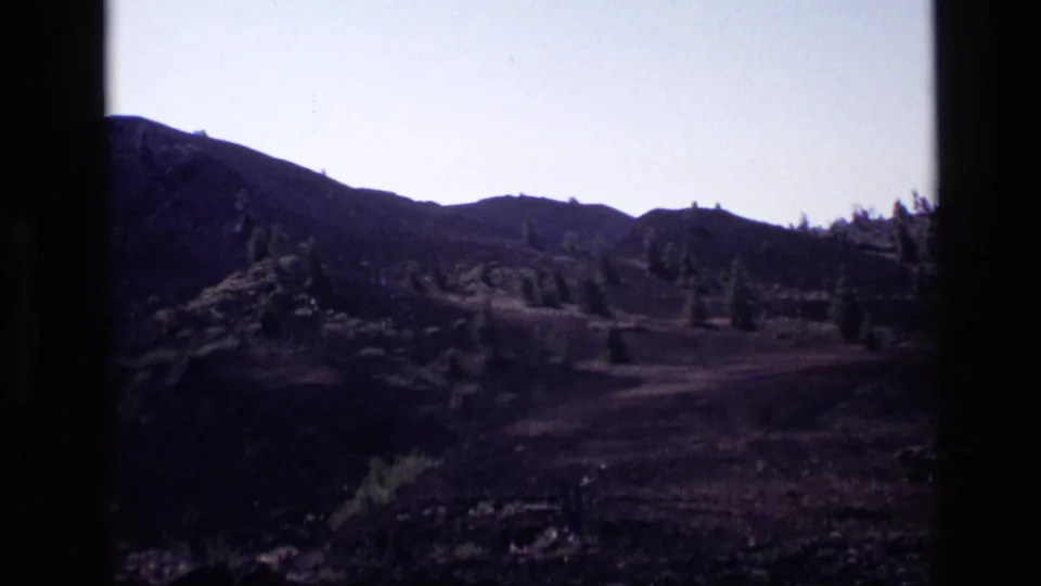 1972 Idaho home-movie frame showing dry mountain terrain, scattered evergreens, and a dark ridge line beneath a pale sky.