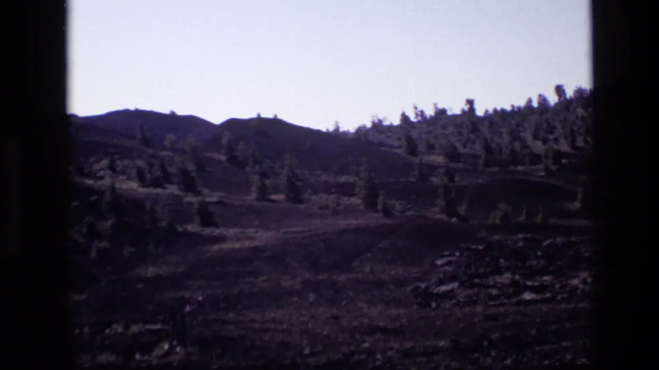 1972 Idaho 8mm frame of rolling rocky ground, scattered evergreens, and low mountain ridges in a quiet landscape view.
