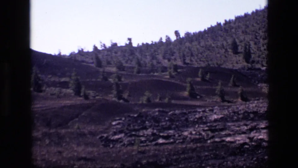 1972 Idaho home-movie view of an evergreen-covered slope, rocky foreground, and pale sky, partly obscured by a dark frame edge.