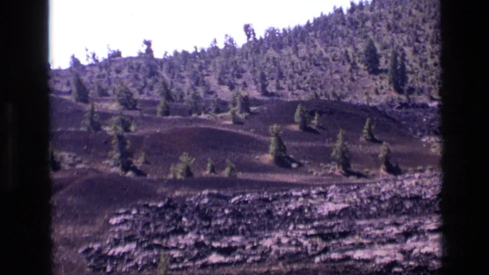 1972 Idaho 8mm frame looking across dark volcanic-looking terrain with small evergreens and a wooded upper slope.
