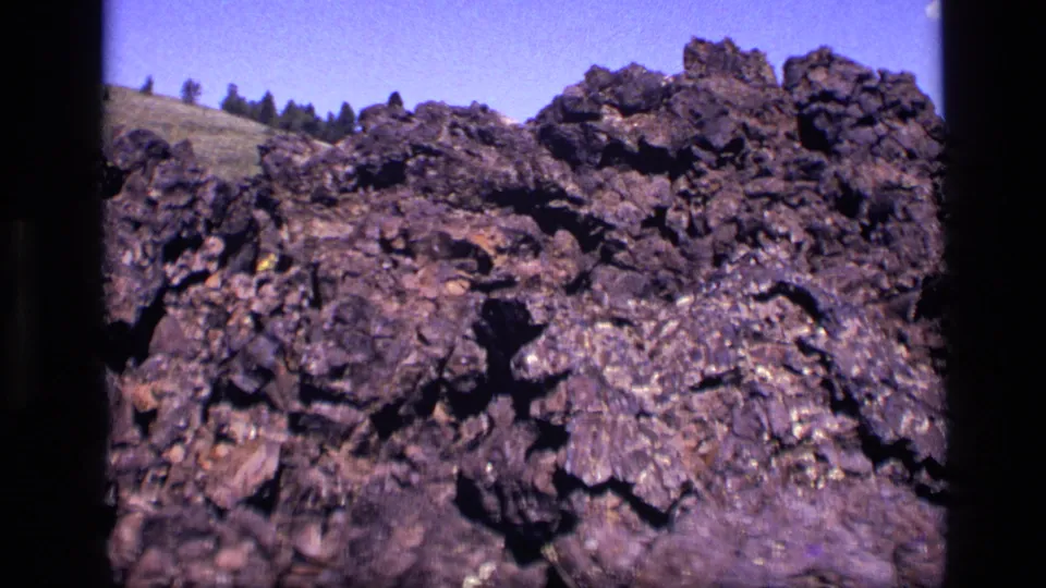 1972 Idaho 8mm frame focused on jagged dark rock formations, with a sparse hillside and blue sky in the background.