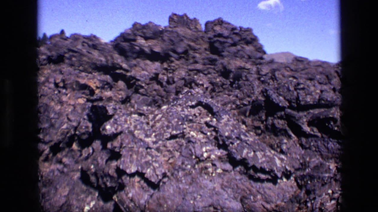 1972 Idaho 8mm frame with a rugged rock ridge, distant hills, and a small cloud in blue sky.