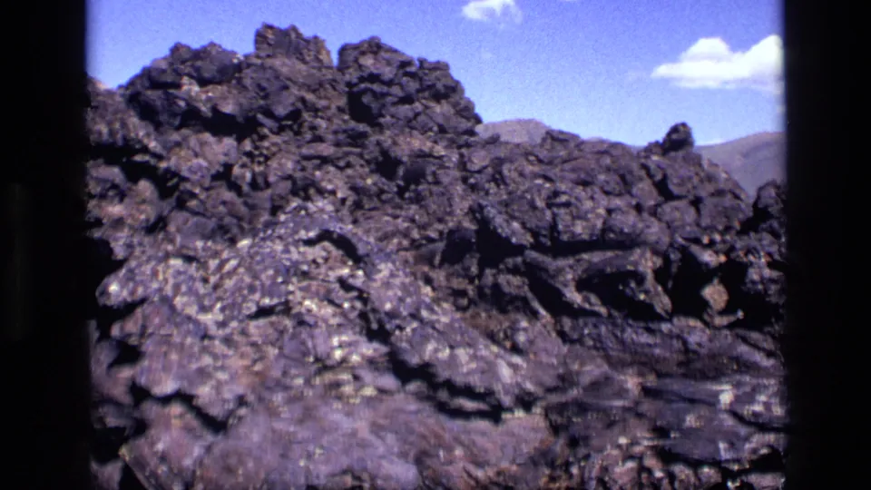 1972 Idaho 8mm frame showing layered dark rocks, distant hills, and open blue sky.