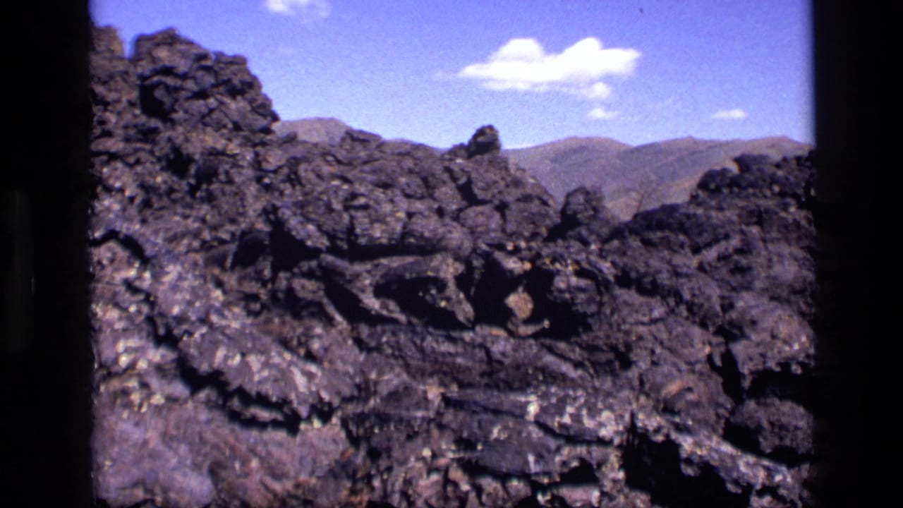 1972 Idaho 8mm frame of rough dark lava rock formations with mountains on the horizon and small white clouds overhead.
