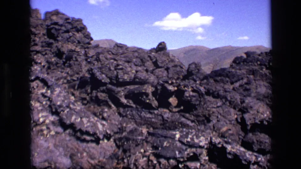 1972 Idaho 8mm frame of rough dark lava rock formations with mountains on the horizon and small white clouds overhead.