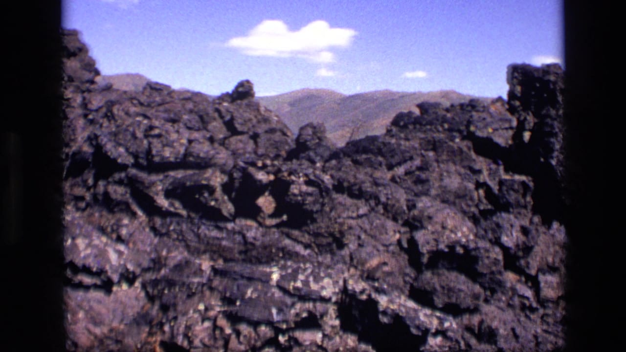 1972 Idaho home-movie view across craggy volcanic rock toward muted mountain ridges beneath a bright blue sky.