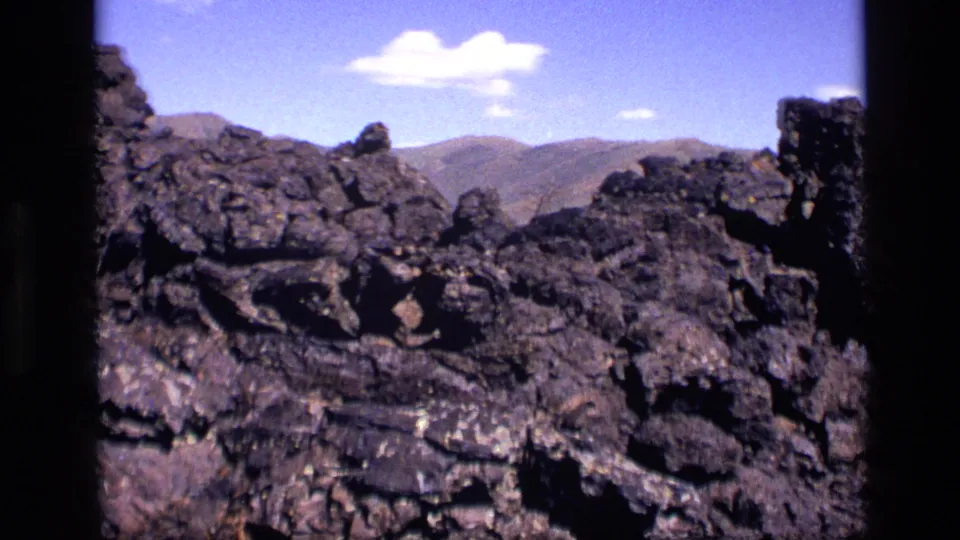 1972 Idaho home-movie view across craggy volcanic rock toward muted mountain ridges beneath a bright blue sky.