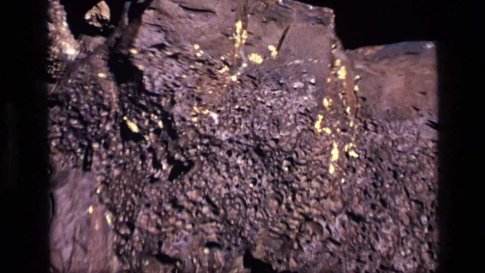 1972 Idaho 8mm frame showing a closer view of rough mineral texture with scattered yellow flecks and heavy shadow around the rock.