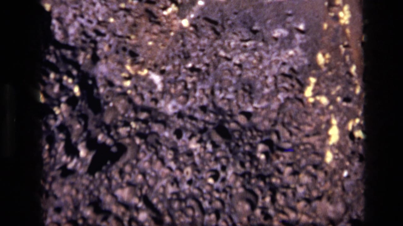 1972 Idaho 8mm close-up of dark porous rock, with pitted texture, black openings, and small reflective flecks across the surface.