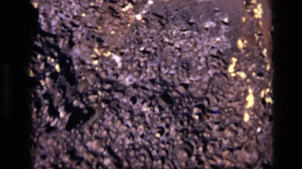 1972 Idaho 8mm close-up of dark porous rock, with pitted texture, black openings, and small reflective flecks across the surface.