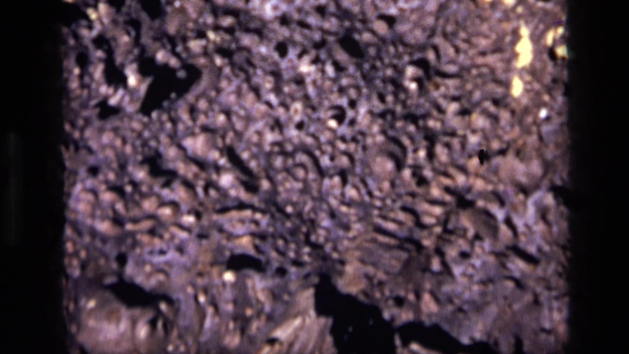 1972 Idaho 8mm frame showing rough volcanic-looking rock, mottled purple-brown color, black crevices, and scattered bright specks.