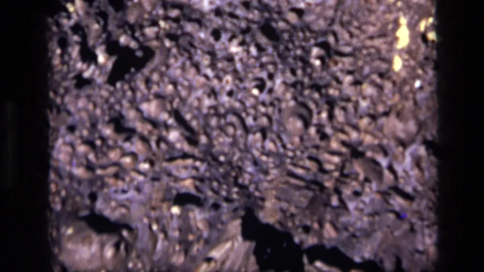 1972 Idaho 8mm close-up of dark porous rock with dense pits, purple-brown cast, and a larger shadowed opening near the bottom.