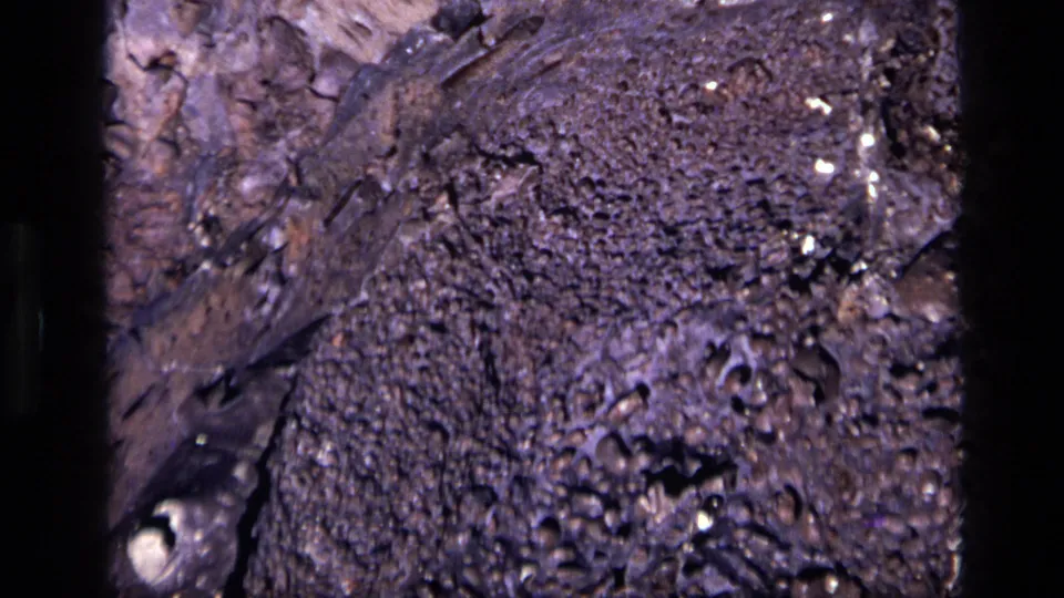 1972 Idaho 8mm frame of a rough, lava-like rock piece with dense pores, dark edges, purple-brown color, and small reflective flecks.