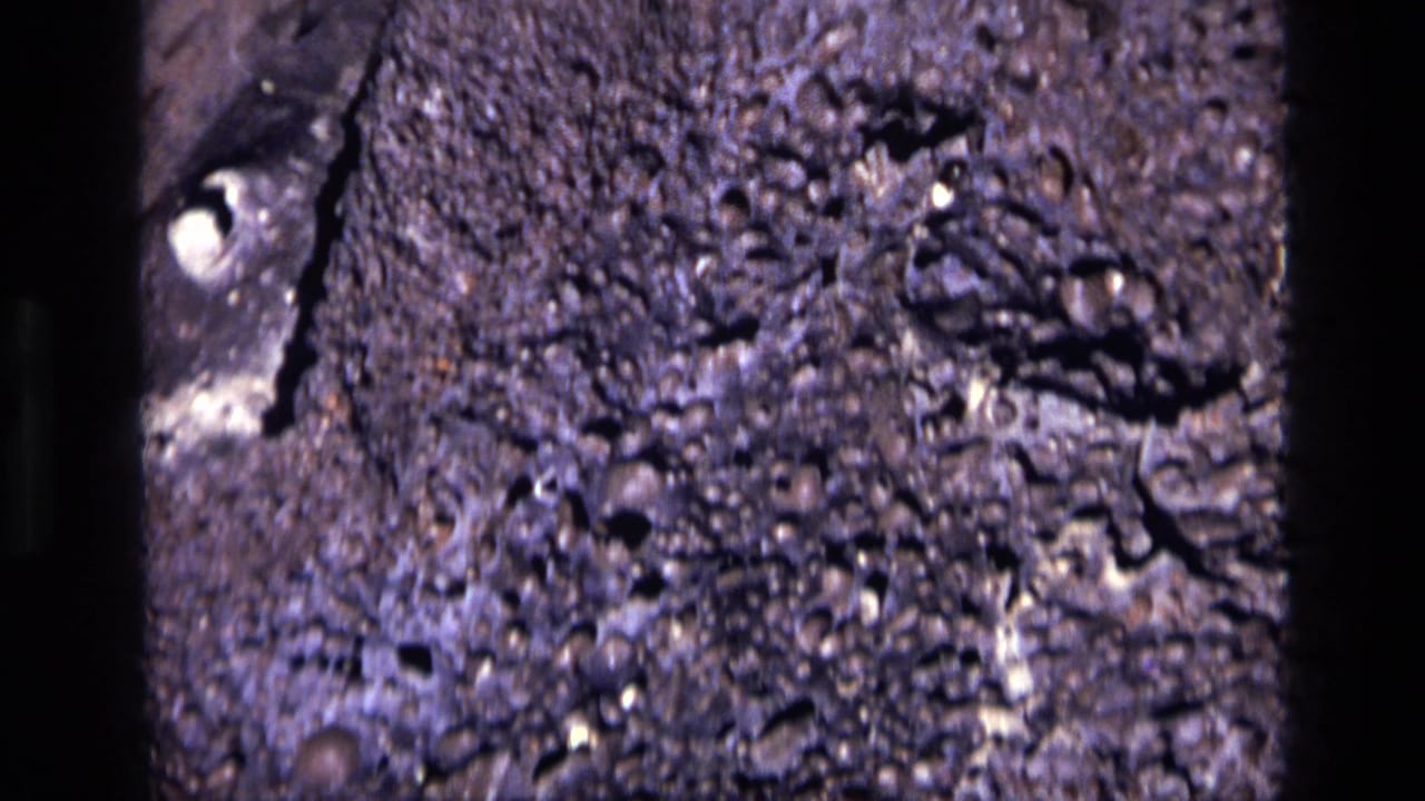Close 1972 Idaho home-movie view of pitted gray rock, showing pale mineral marks and irregular shadowed holes.