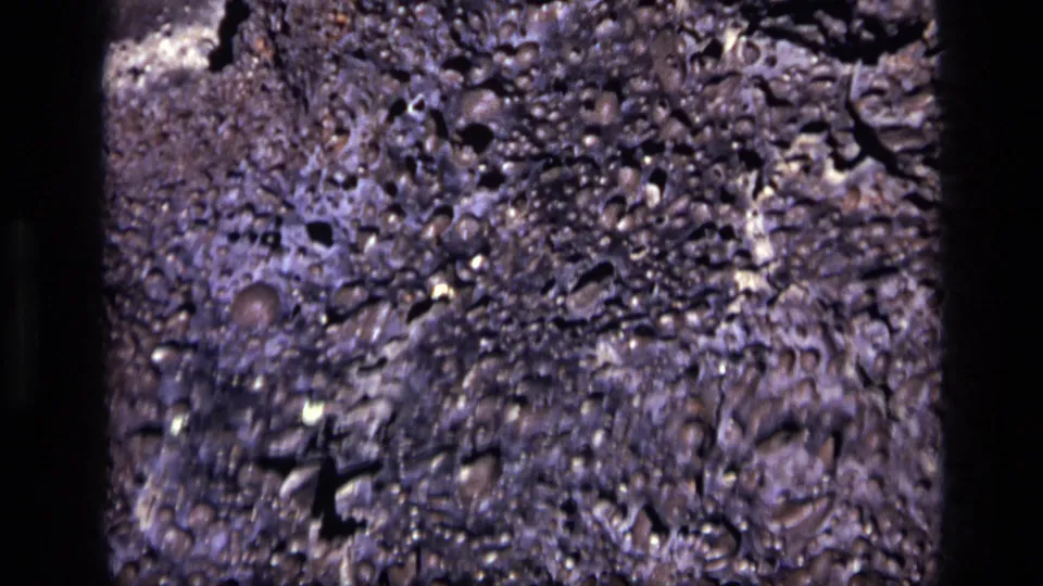 Idaho 1972 8mm frame of dense gray rock texture with small pits, white mineral flecks, and dark edge shadowing.