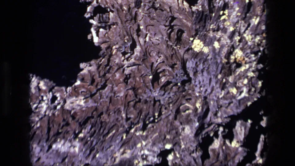 1972 Idaho 8mm frame showing ridged dried material, black voids, and bright flecks across a rough textured surface.